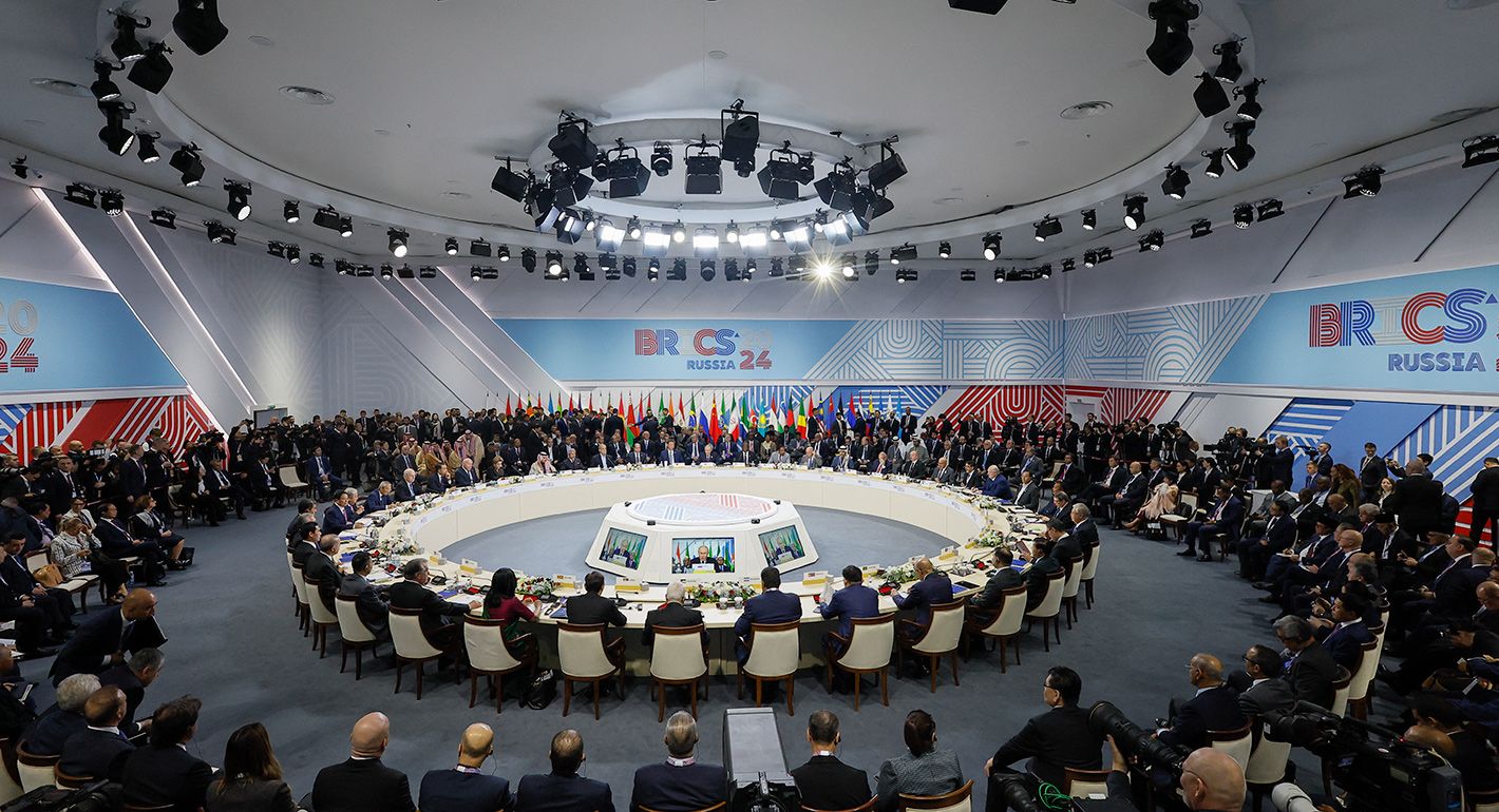 Delegates seated around the round table at the BRICS 2024 summit in Russia, with national flags in the background.
